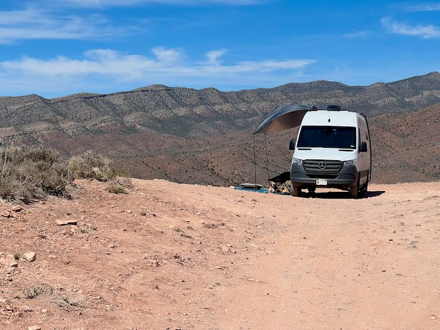 Explore the desert in comfort with an RV rental. A white camper van with an awning faces arid mountains under a blue sky.