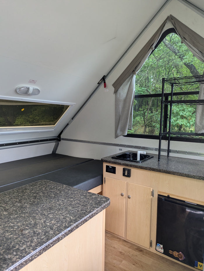 Interior of a compact rv rental, showing a bed, kitchen counter with sink, and a leafy outdoor view through the window.