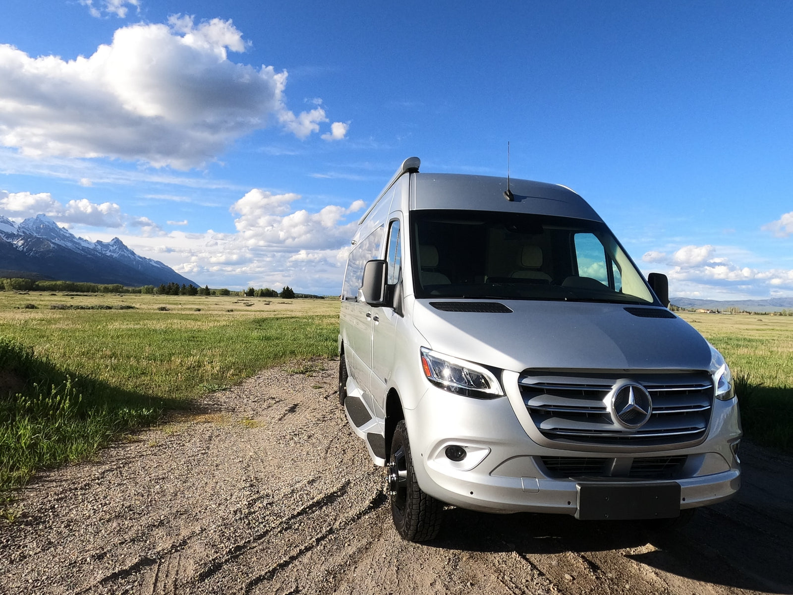 A silver Mercedes Sprinter RV rental van on a scenic gravel road, with snow-capped mountains & blue sky. Ideal for adventure.