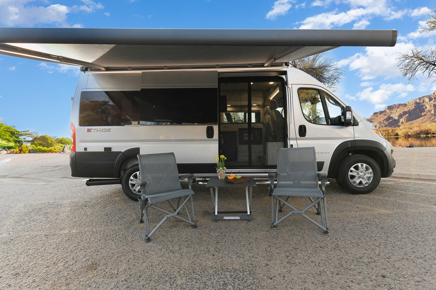 Ethos RV rental: silver camper van w/ extended awning, chairs & table by a scenic river & mountains.