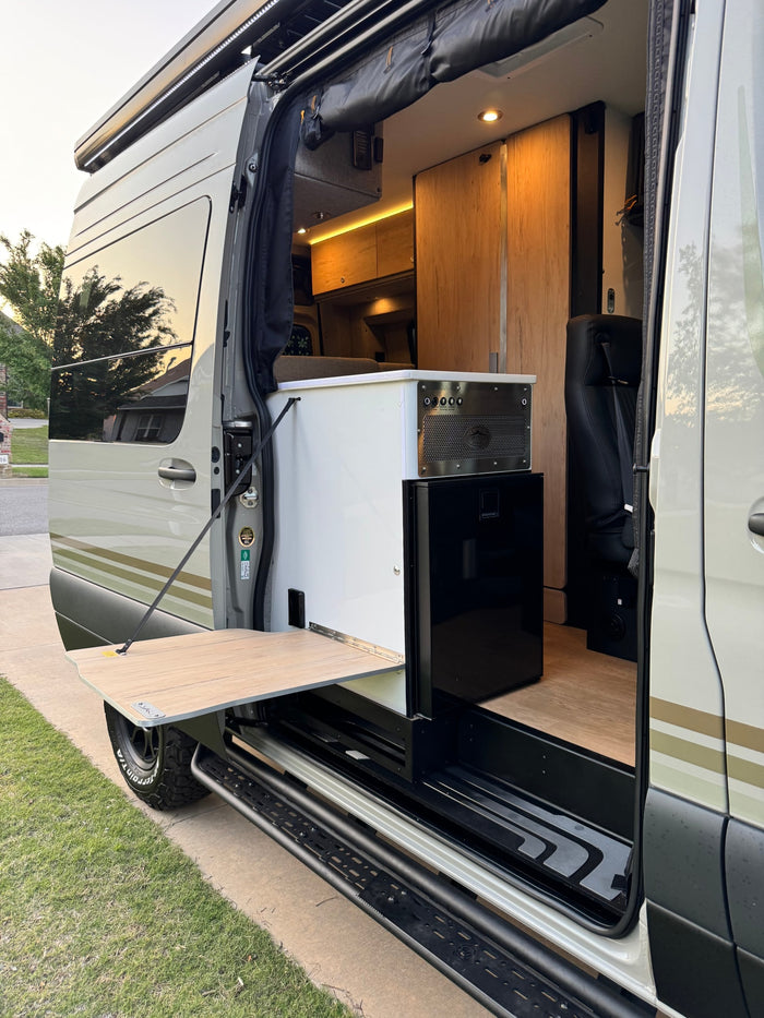 Inside this RV rental: open side door view of a compact kitchenette, fridge, wood cabinets, and an exterior fold-out table.