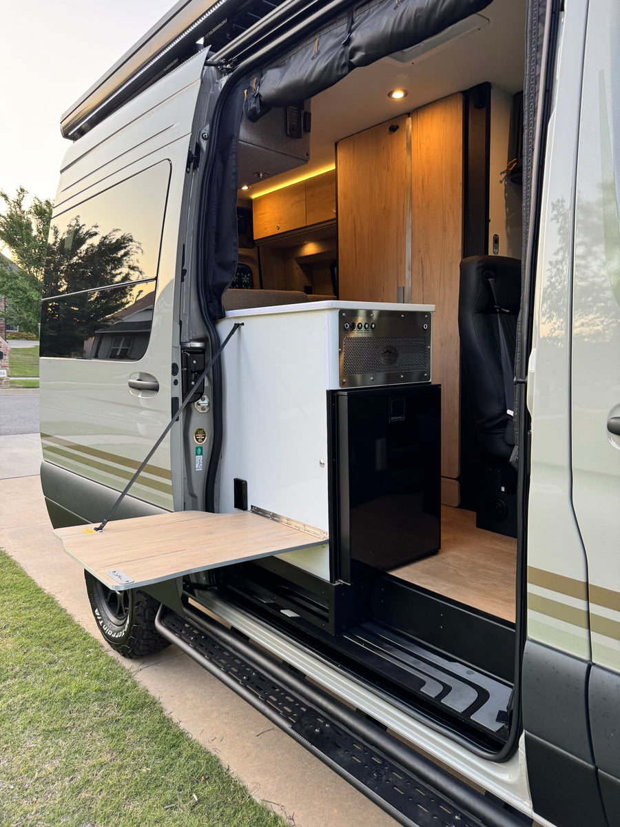 Inside this RV rental: open side door view of a compact kitchenette, fridge, wood cabinets, and an exterior fold-out table.