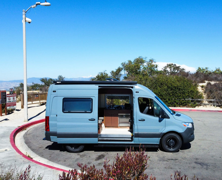 This blue camper van rv rental, with its side door open revealing a modern interior kitchen, is parked at a scenic lookout.