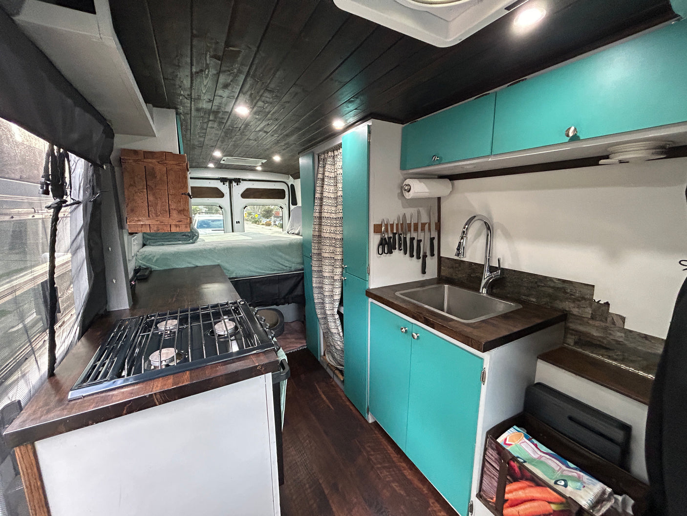 RV rental: Custom van interior with functional kitchen, teal cabinets, cozy bed, and rustic wood finishes for van life.
