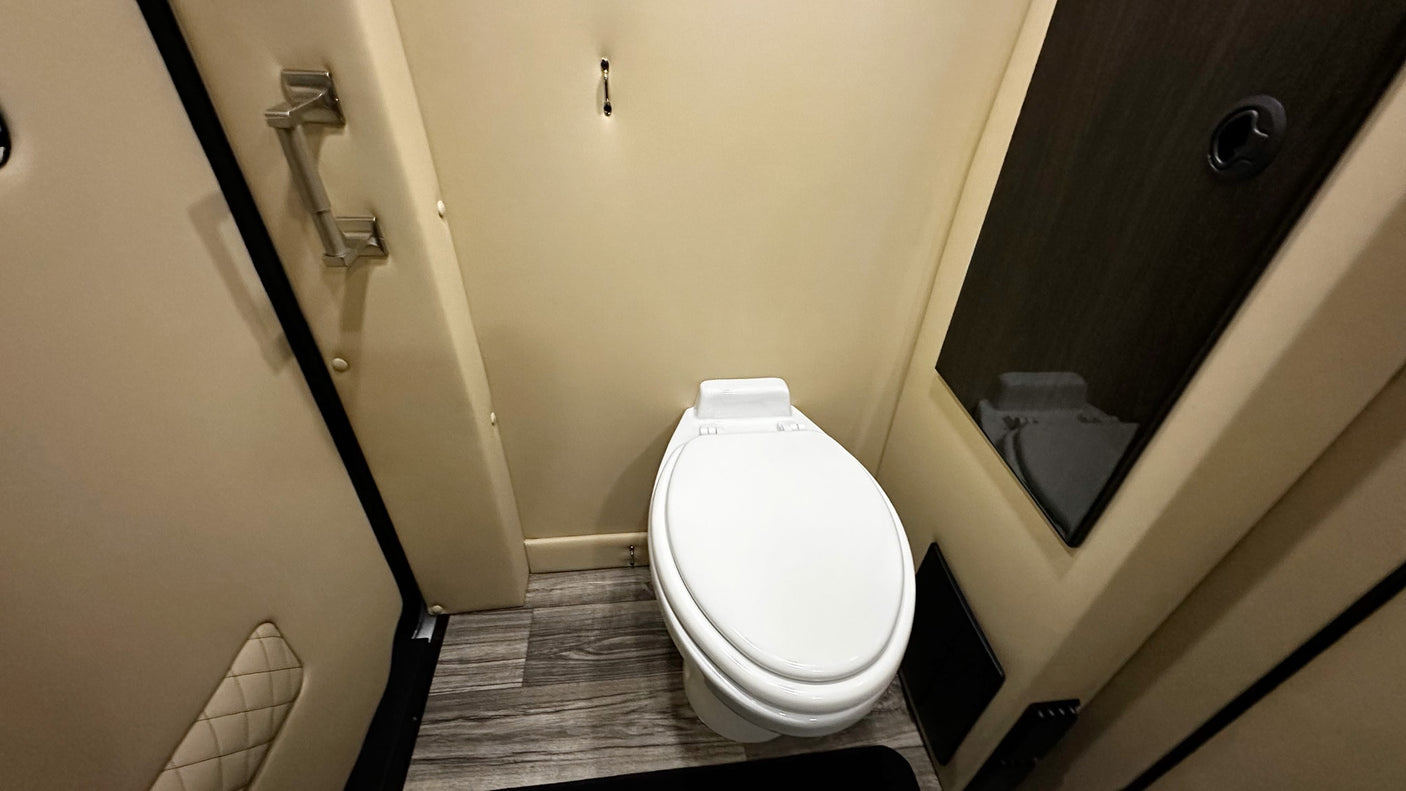 Compact RV rental bathroom features white toilet, padded beige walls, wood-look floor, and dark cabinet.