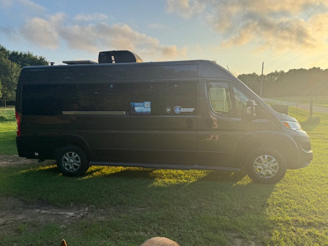 Black RV rental van with "DAZZLE" & "HUMIDITY" logos, parked on grass bathed in golden hour sunlight.