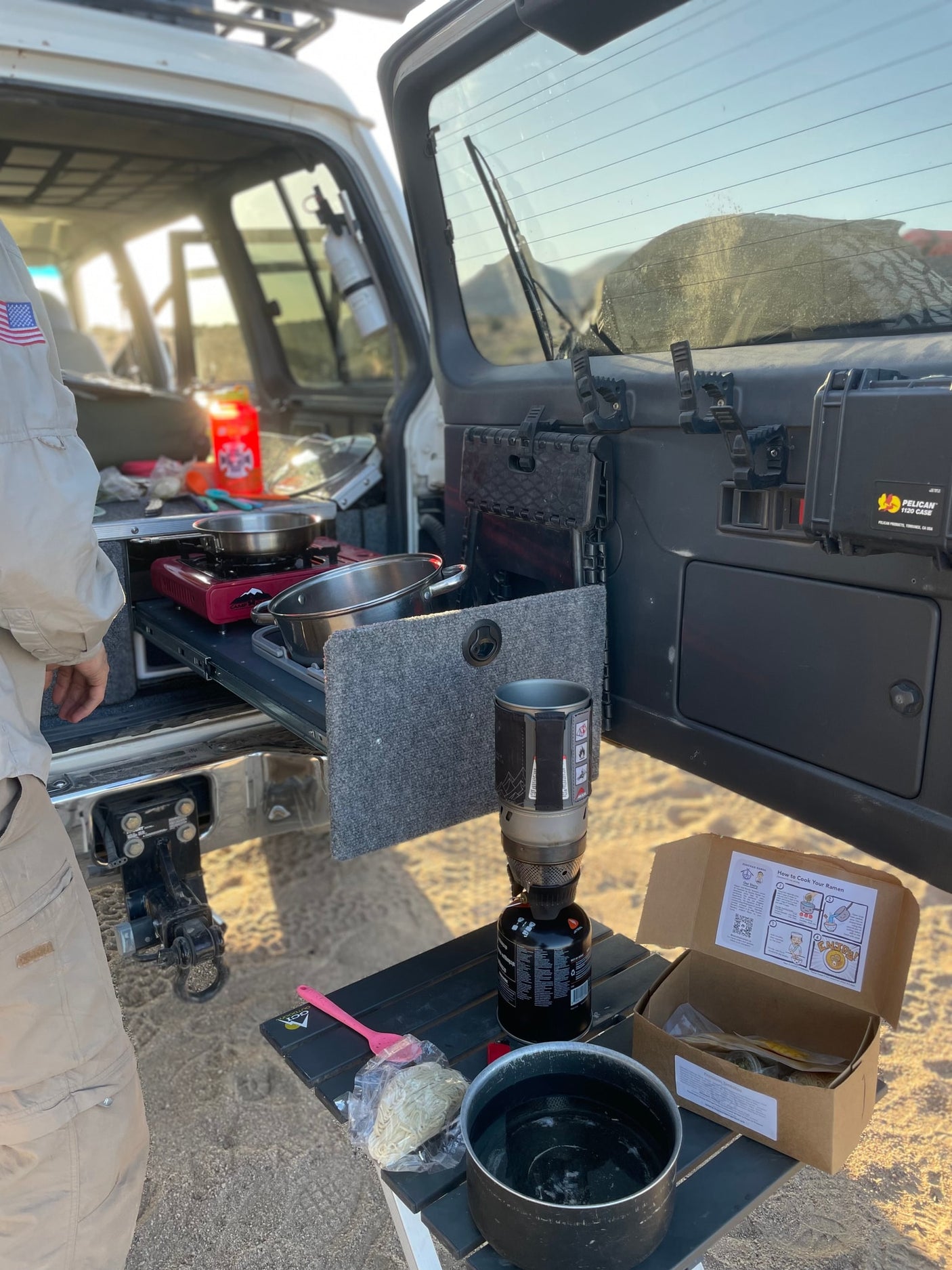 An RV rental's overland camp kitchen setup: cooking ramen with portable stoves and vehicle-mounted gear for an outdoor meal.