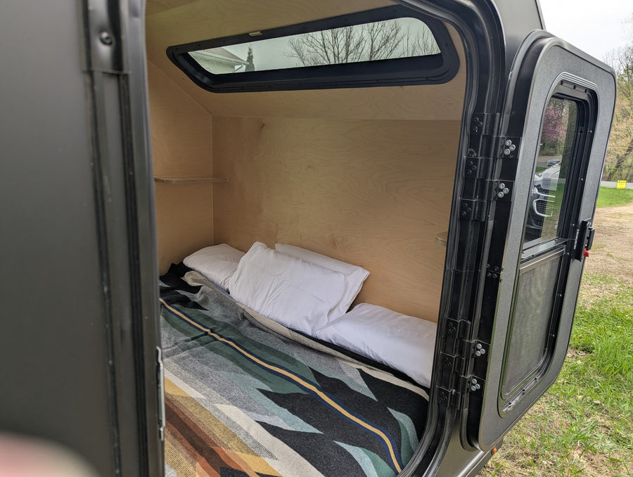 Compact RV rental: A cozy teardrop trailer bedroom with a patterned blanket, white pillows, window, and open door view.