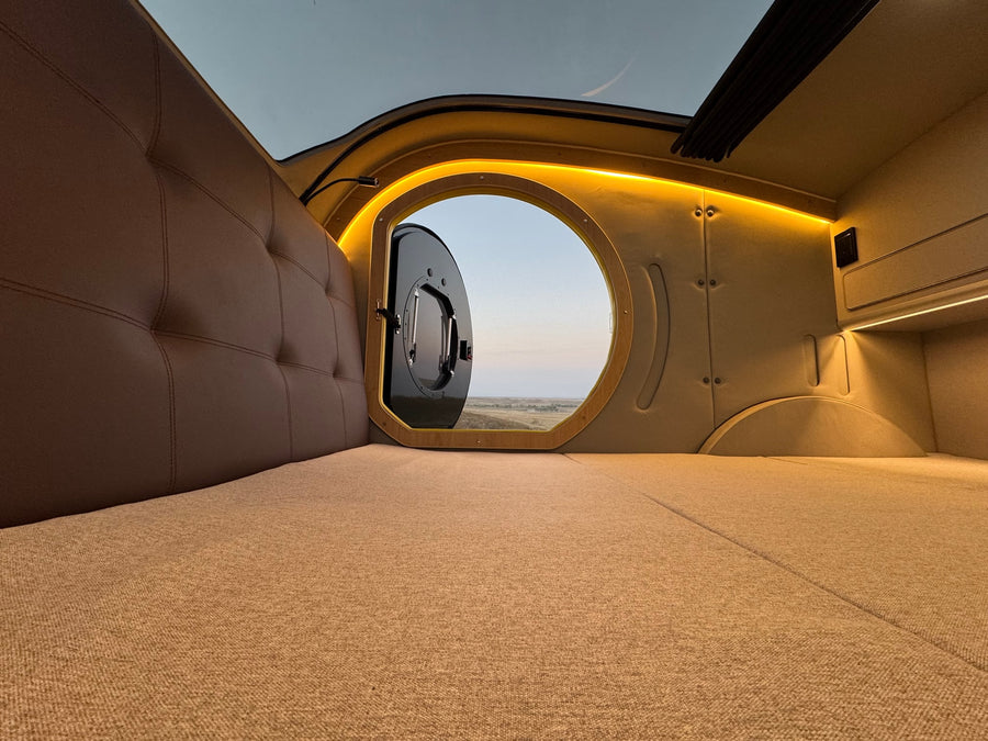 Cozy RV rental interior with a comfortable bed and scenic porthole view of the landscape.