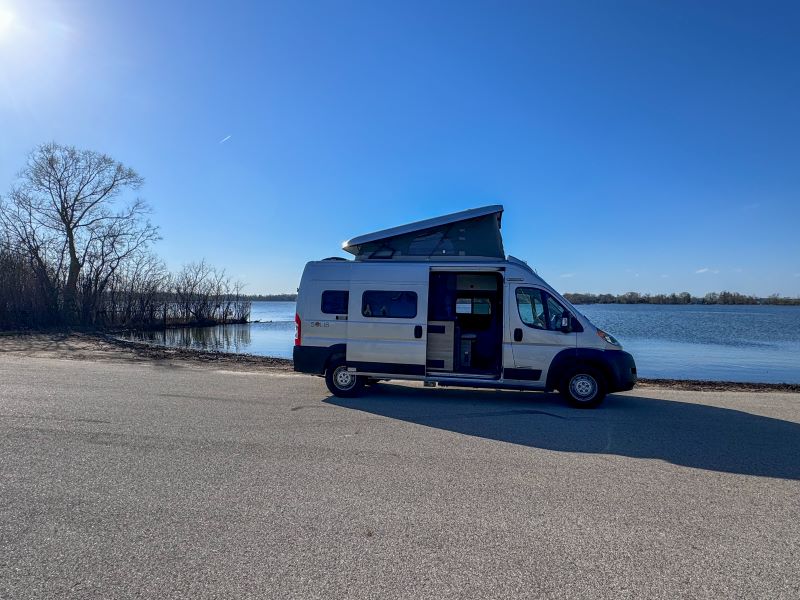 Scenic RV rental: a silver camper van with pop-top open, parked on asphalt beside a lake under a clear blue sky.