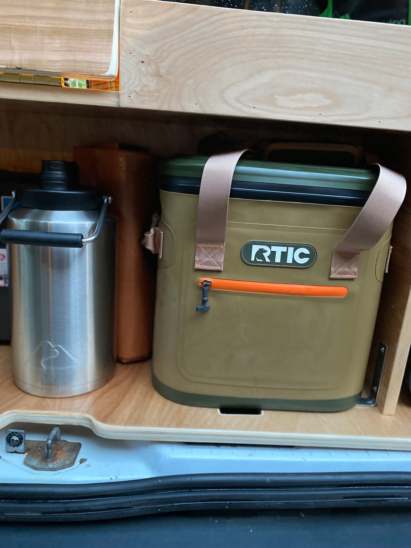 RV rental setup: olive RTIC cooler & silver mountain-logo water bottle on custom wood storage shelves.