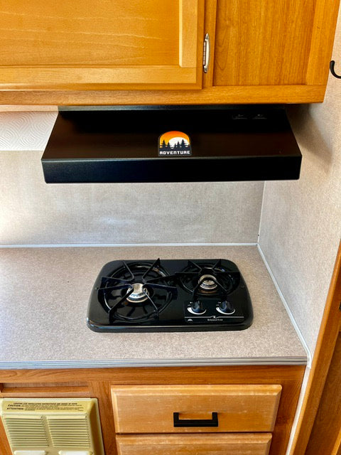 RV rental kitchen with a black two-burner stovetop, range hood with "ROVENTURE" logo, and wood cabinets.
