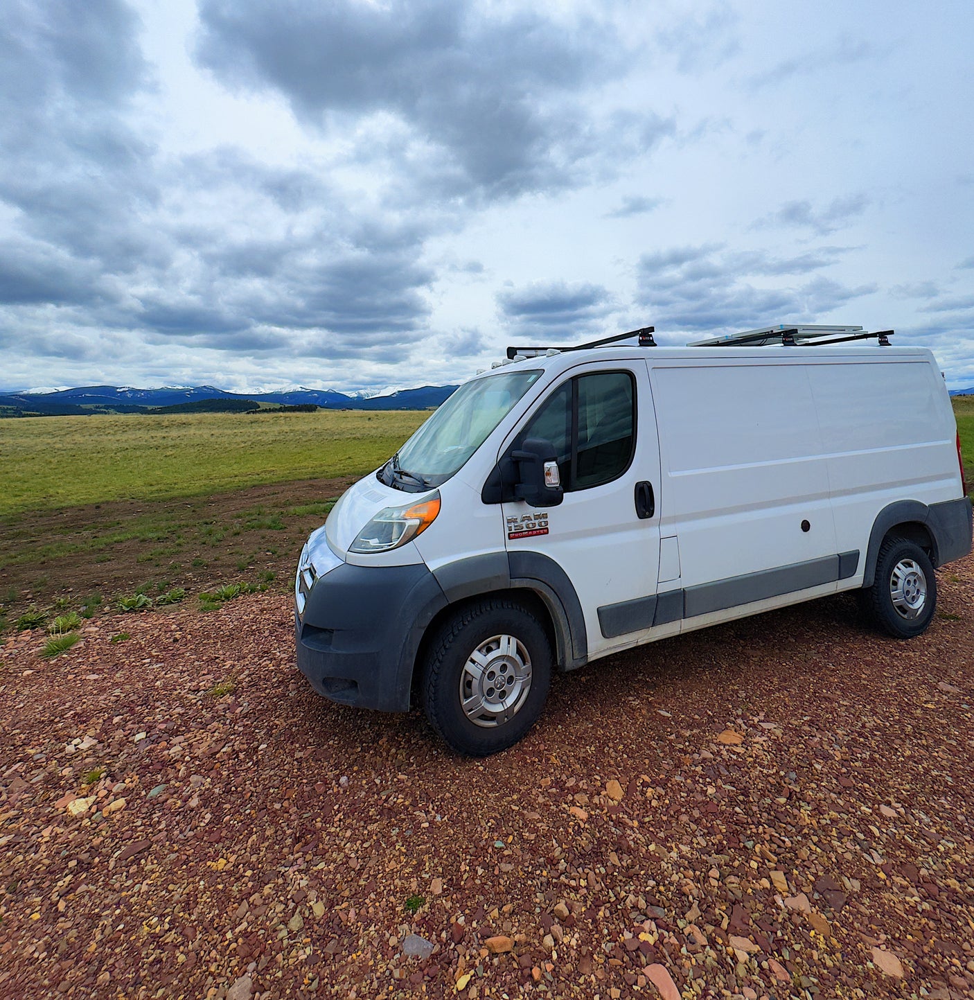 White Ram ProMaster camper van (RV rental) on a gravel road, backed by snowy mountains under cloudy skies. Explore the outdoors!