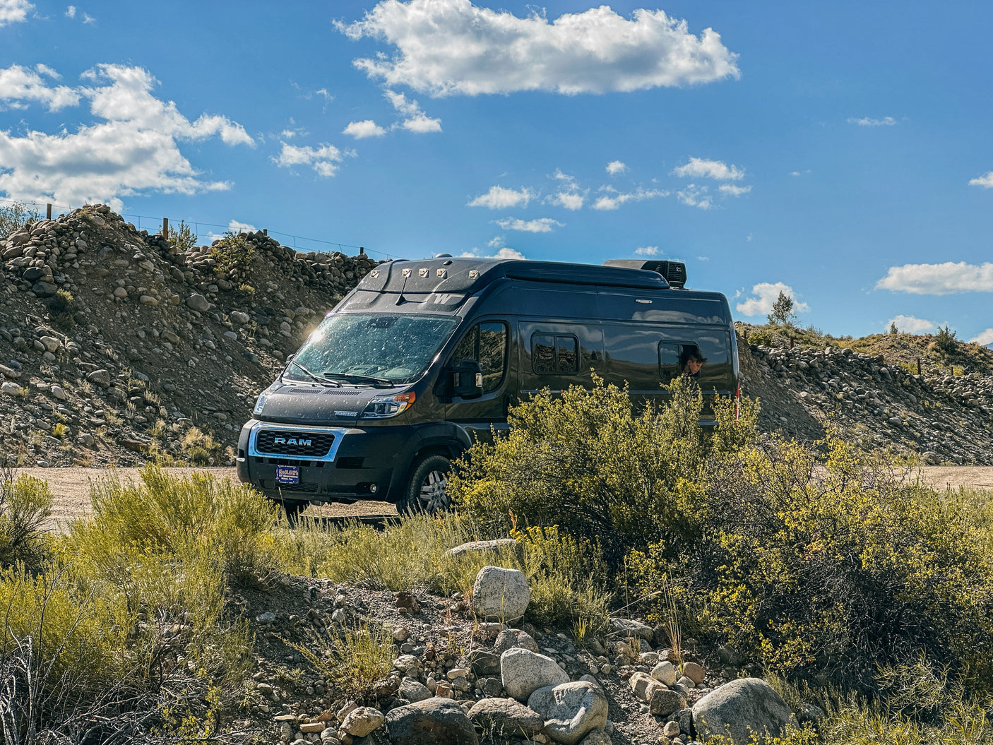 Adventure awaits with this RV rental! Dark camper van parked in a sunny, rocky outdoor setting, person in window.