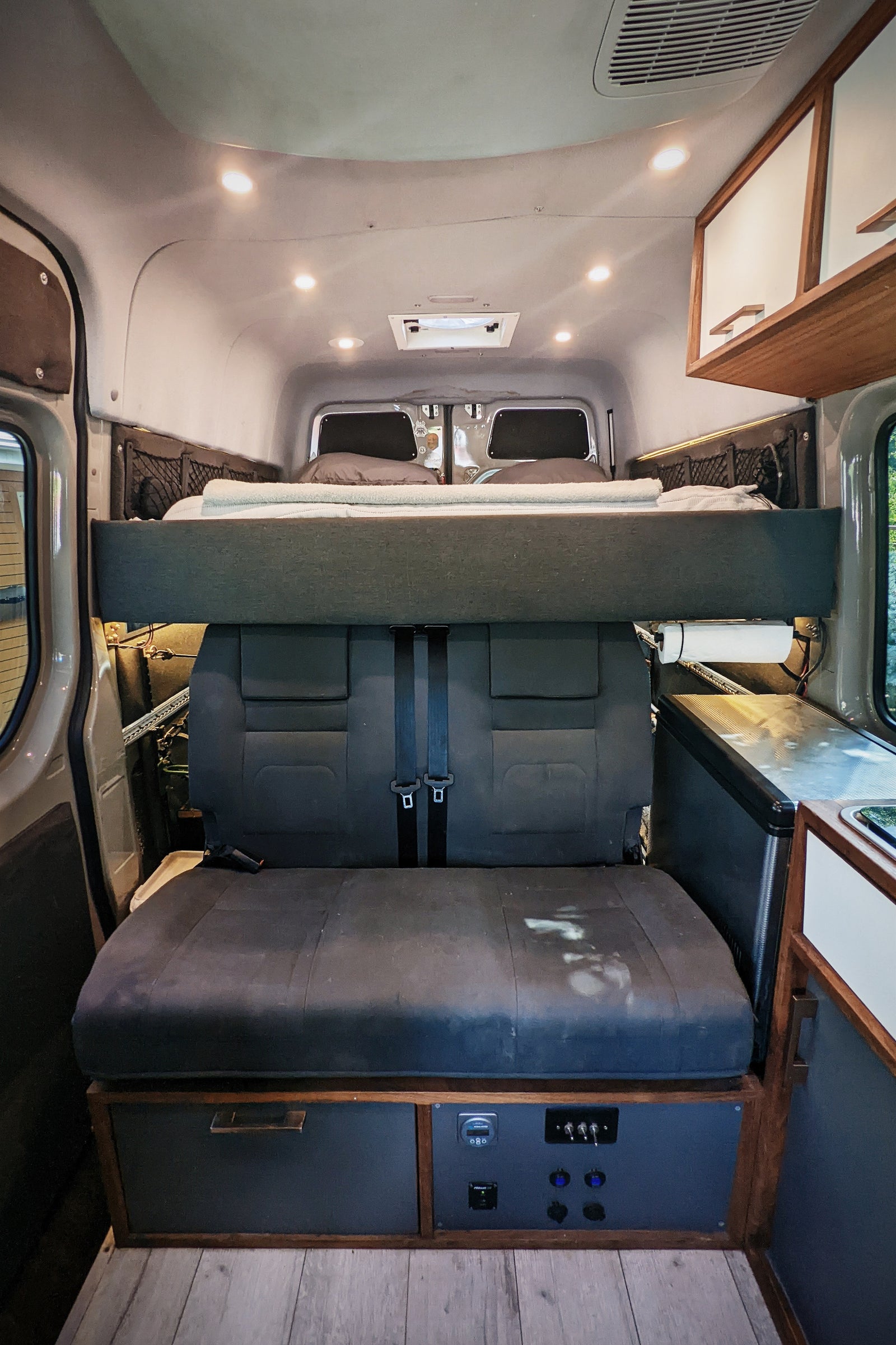 RV rental interior: elevated bed over rear seats, kitchen, and power control panel. Ready for your camper van trip.