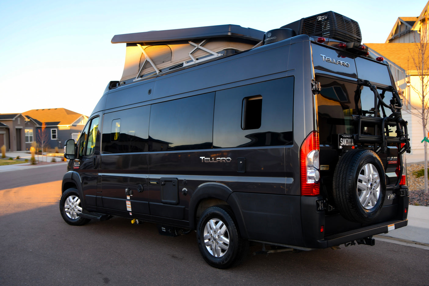 Dark gray Tellaro camper van RV rental parked on a street at sunset, featuring a pop-top roof and a rear bike rack.