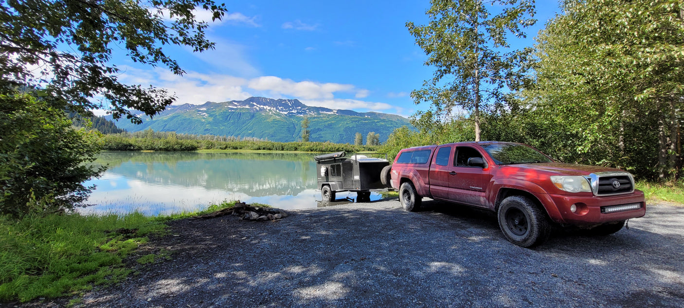 Red truck with an RV rental trailer backed by a serene lake and snow-capped mountains. Ideal for off-grid camping.