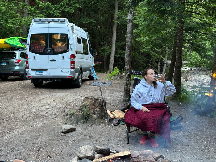 Relaxing by a campfire with an RV rental van and a car carrying kayaks in a lush forest, perfect for outdoor adventure.