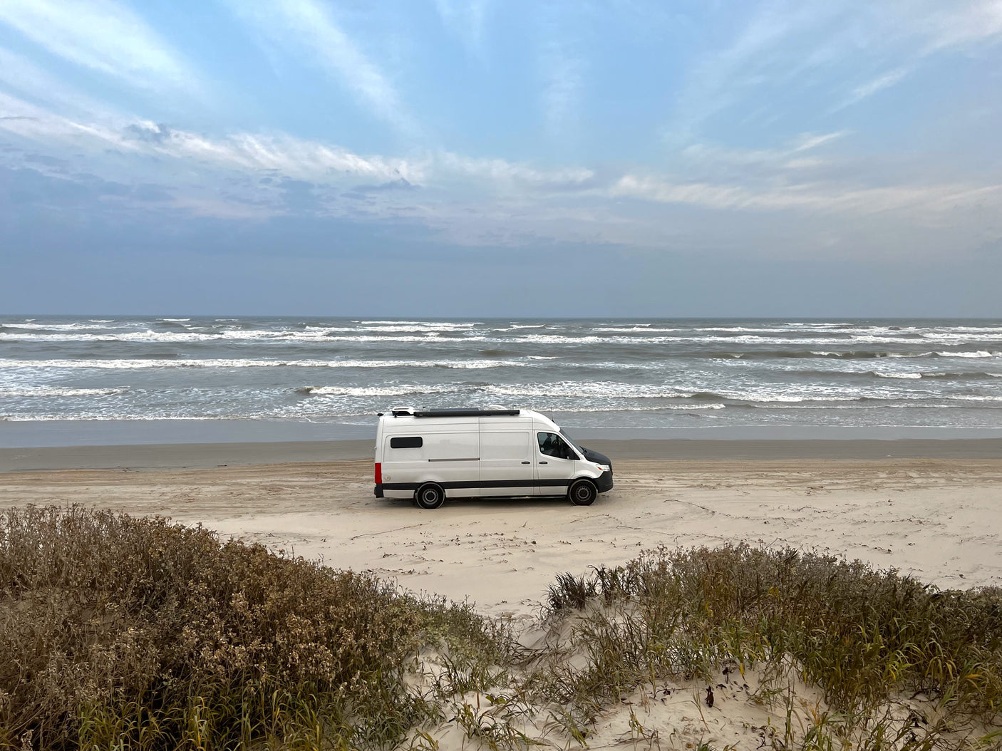 White RV rental camper van parked on a sandy beach. Ocean waves crash under a cloudy sky, ideal for beach camping.