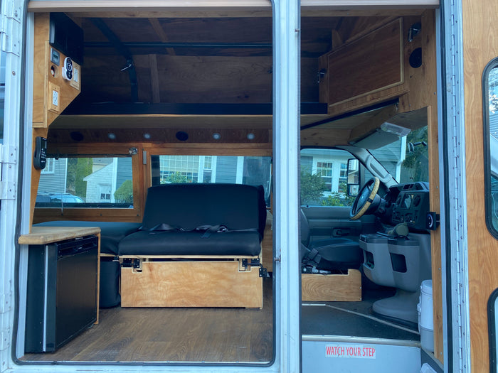 RV rental: Custom camper van interior with warm wood finishes, versatile seating/bed, mini-fridge, and driver's cab.