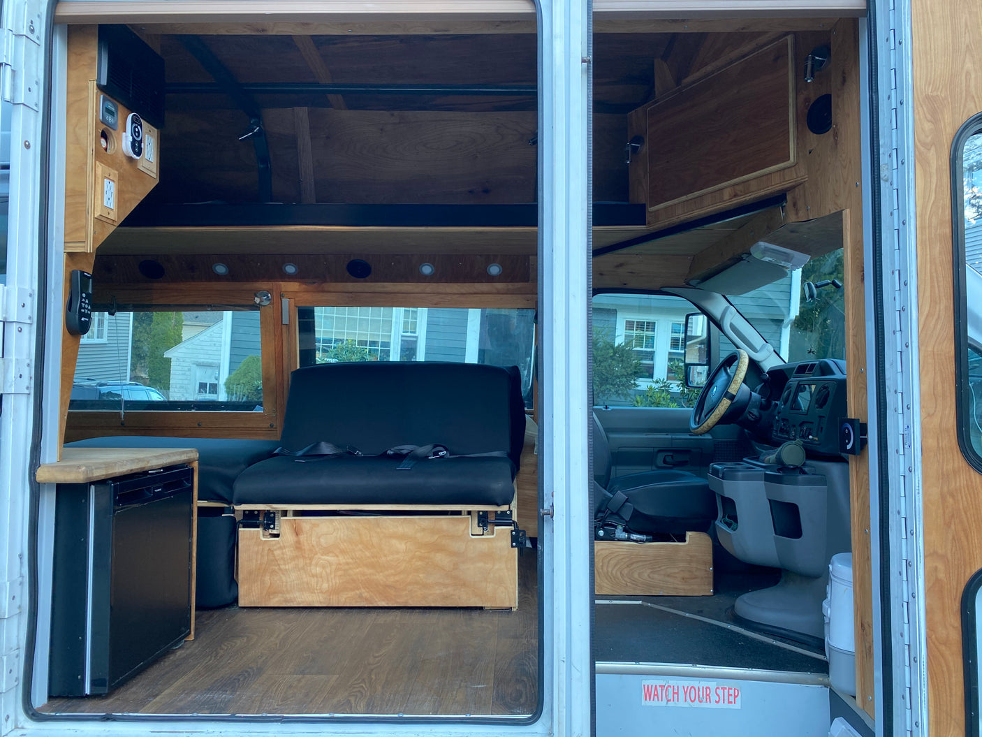 RV rental: Custom camper van interior with warm wood finishes, versatile seating/bed, mini-fridge, and driver's cab.