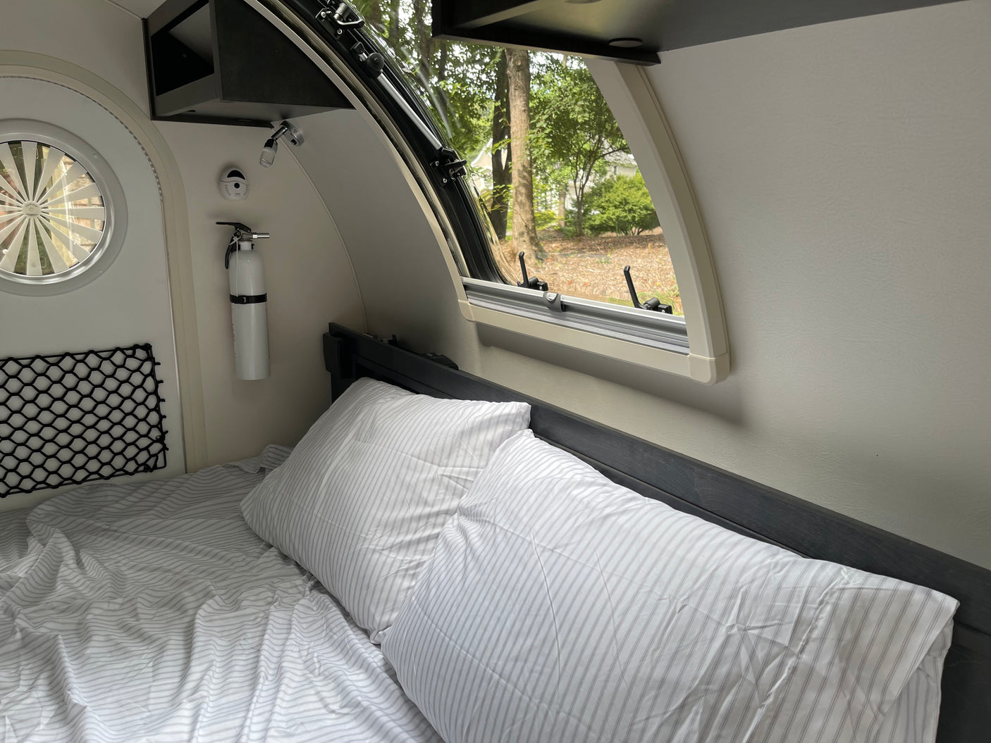 Cozy RV rental interior with a bed, window view of trees, and a fire extinguisher. Ideal for comfortable travel adventures.