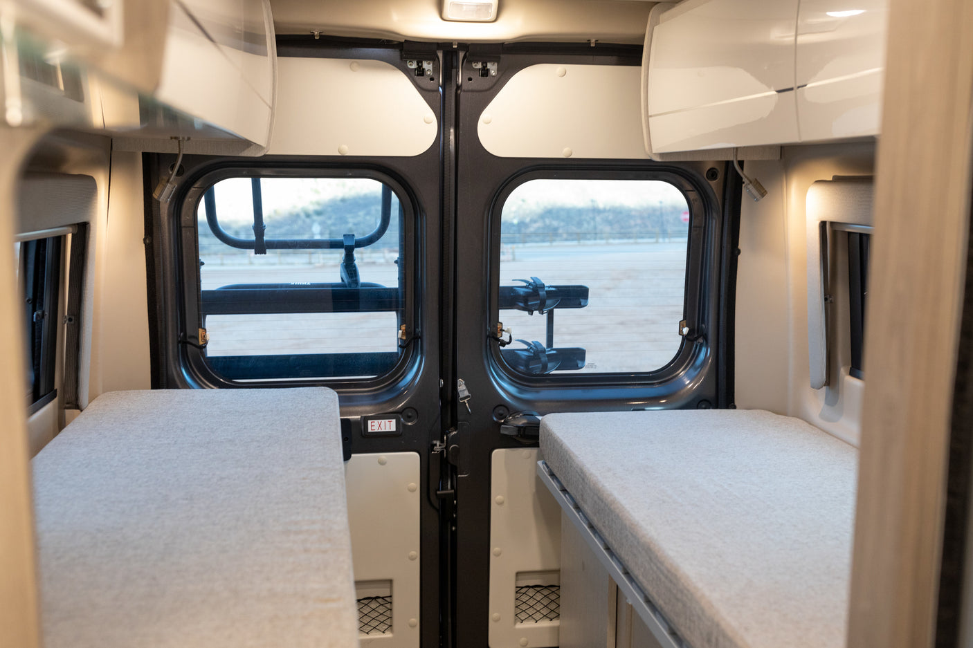 RV rental interior: two cozy beds, overhead storage, and rear doors with windows, including an 'EXIT' sign.