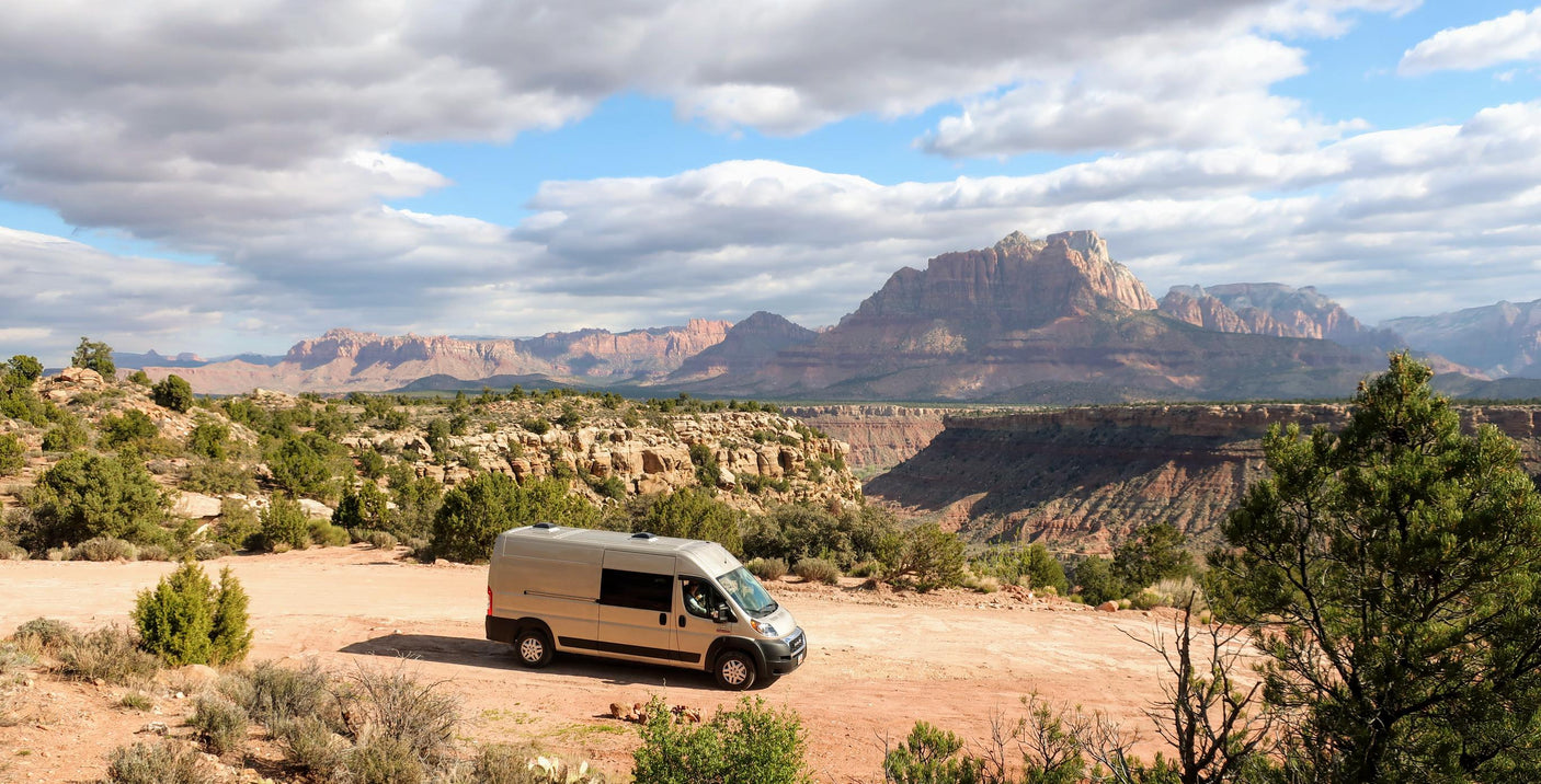Light-colored RV rental on a dirt road in a vast desert landscape with red rock canyons and majestic mountains.