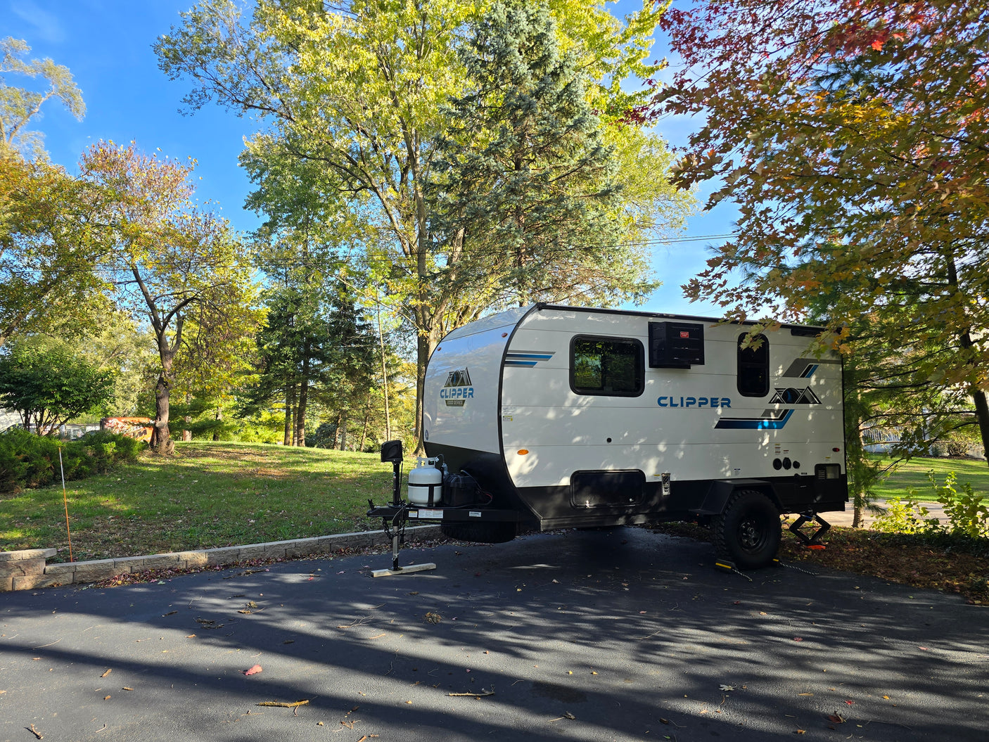White Clipper travel trailer rv rental parked on asphalt, surrounded by bright autumn trees under a clear blue sky.