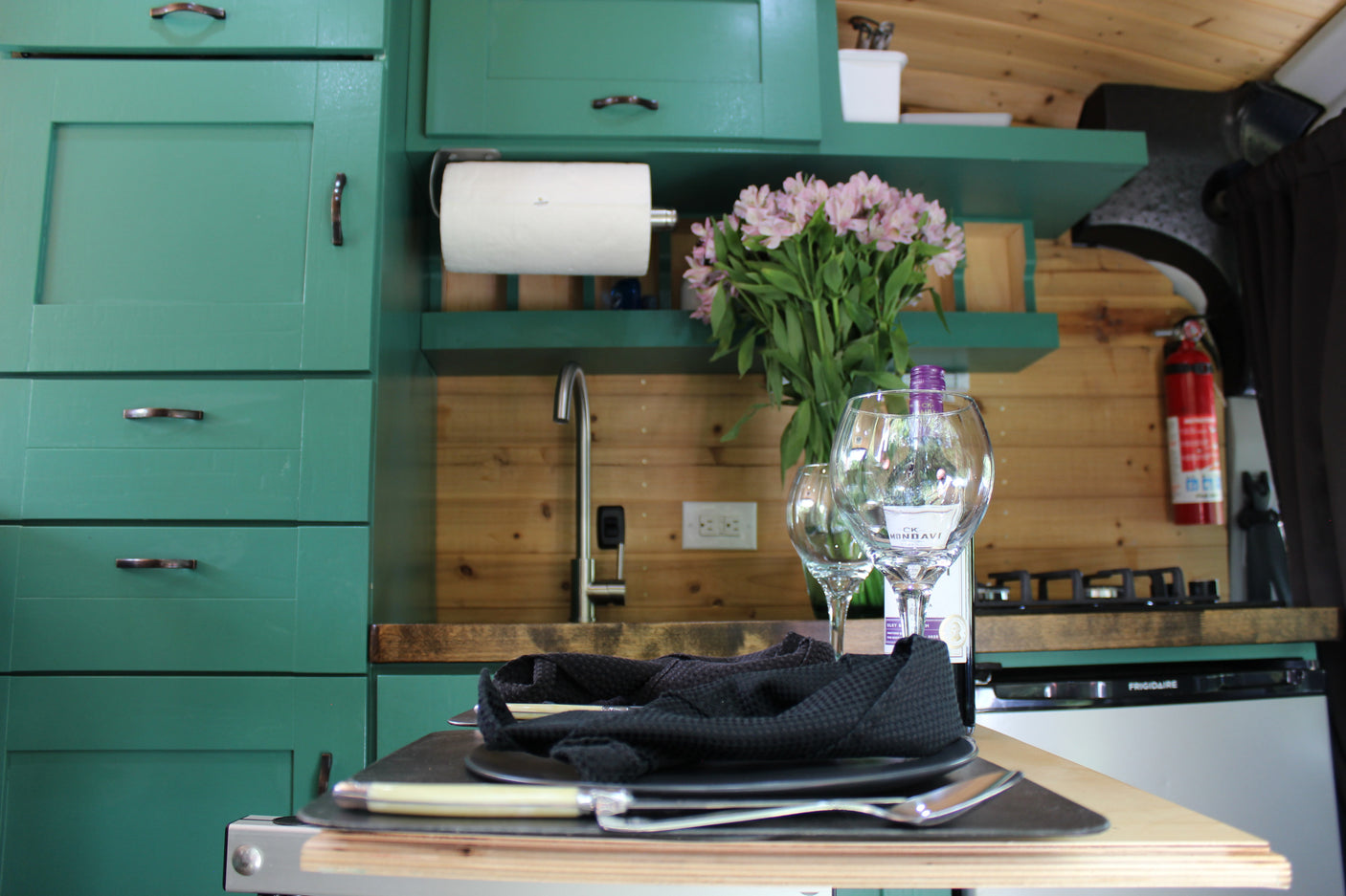 Stylish RV rental kitchen with green cabinets, sink, dining for two, wine, flowers, and essential amenities.