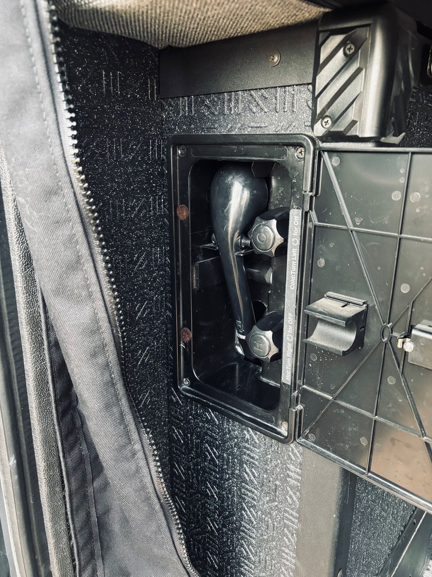 Compact outdoor shower system in an RV rental, showing a black shower head and hot/cold controls inside its compartment.