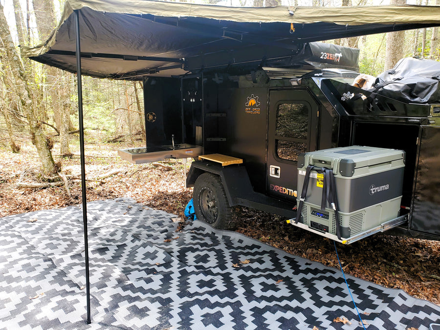 An RV rental overland trailer campsite with outdoor kitchen, Truma fridge, awning, and ground mat. Perfect for off-grid adventure!