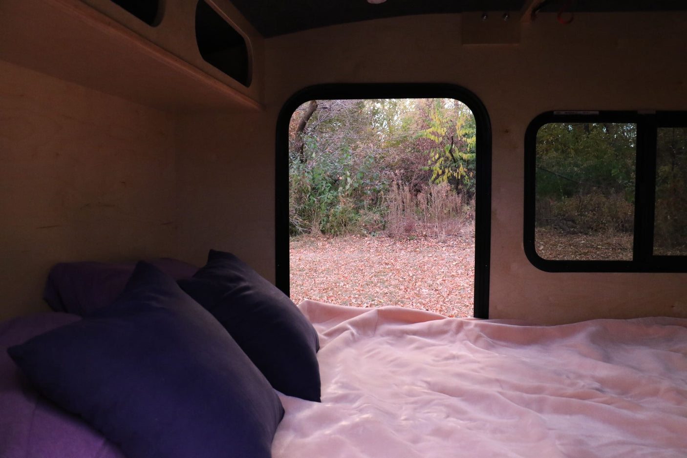 and windows looking out to autumn trees. Camper living.|Inside a blue rv rental: a cozy bed with pillows looks out to a scene of autumn leaves on the ground.|Cozy RV rental interior features a bed with pillows & a window view of fall foliage.|Inside an RV rental: a cozy bed with purple pillows and a view of autumn leaves through two windows.|Cozy RV rental interior: light wood paneling