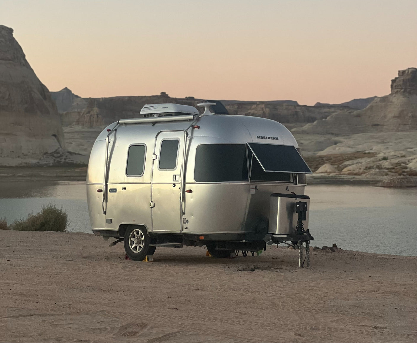 An Airstream RV rental camps by a serene lake at dusk, surrounded by rugged desert mountains, offering a perfect outdoor escape.