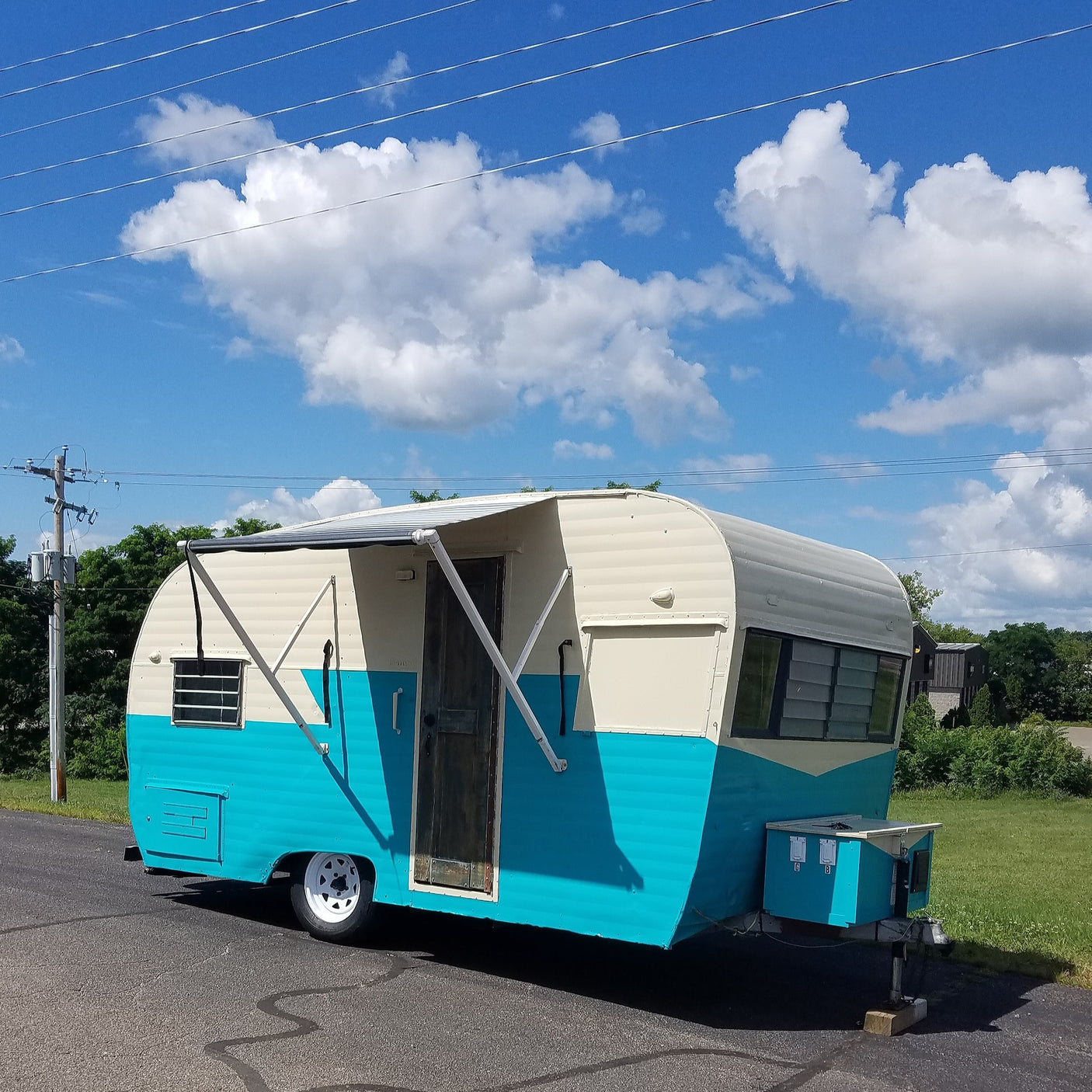 A retro turquoise and cream RV rental, featuring an extended awning, sits on asphalt under a vibrant blue sky with clouds.
