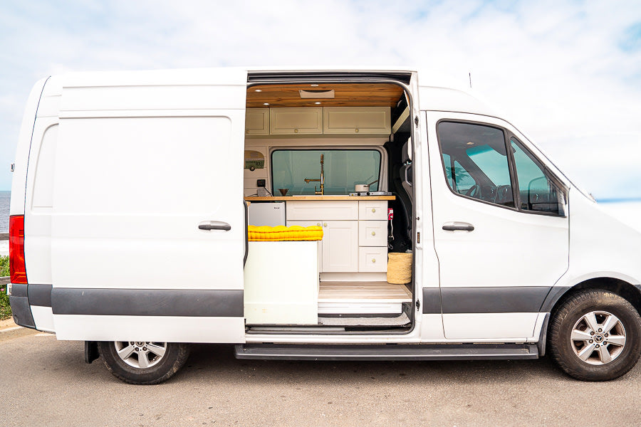 This custom white camper van, perfect for an RV rental, shows its open side door revealing a stylish kitchen and cozy interior setup for travel.