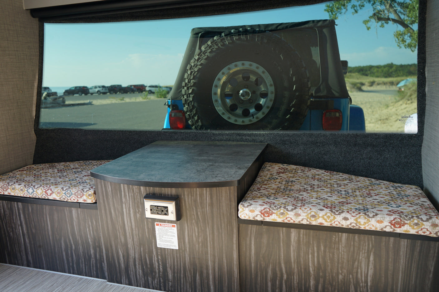 A cozy RV rental interior with patterned cushions, a table, and a window view of a blue Jeep parked near a beach.