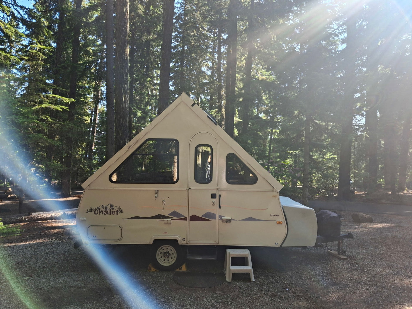 A beige A-frame pop-up RV rental is parked on gravel at a sunny forest campsite