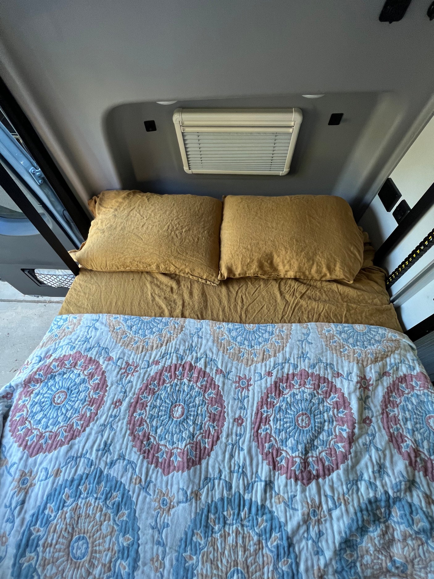 The sleeping area of an RV rental, featuring a bed with mustard pillows, a patterned quilt, and a ceiling vent.