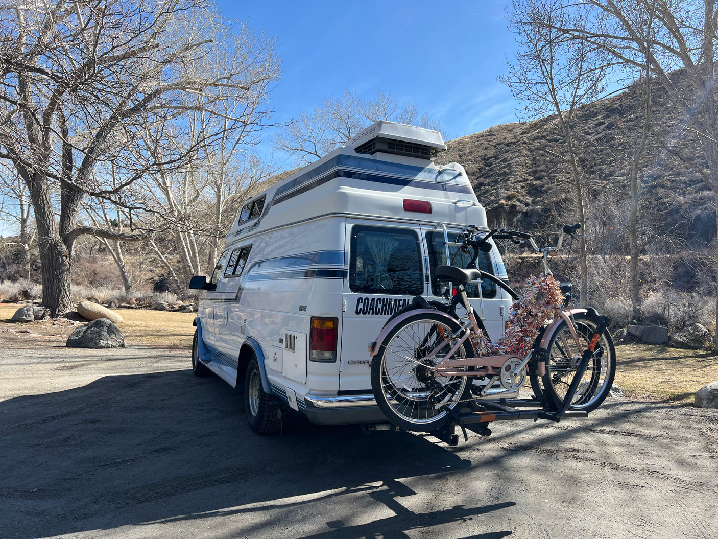 White Coachmen RV rental with two bikes on a rack, parked among bare trees and a hillside on a sunny day.