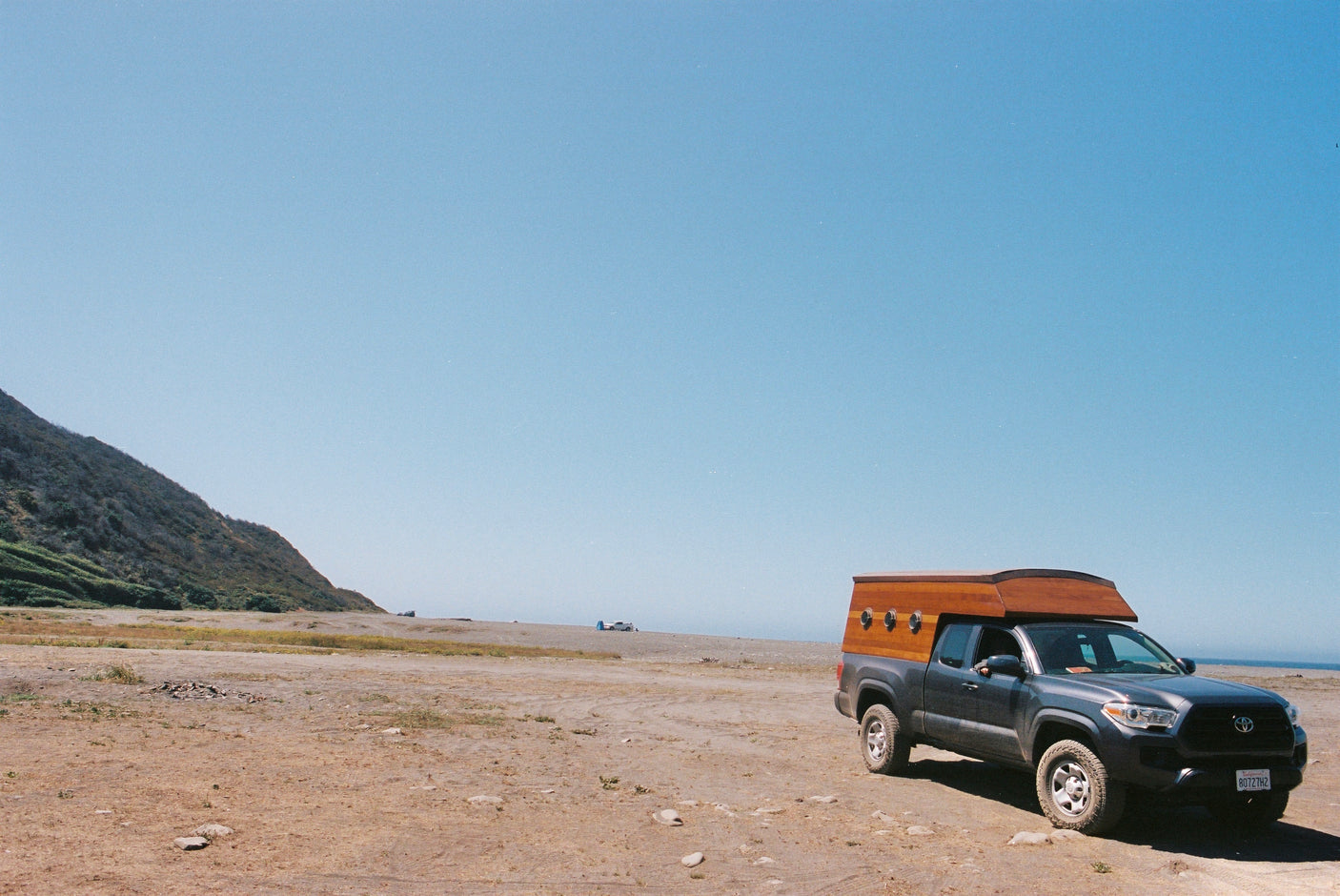 A gray Toyota Tacoma truck with a unique wood RV rental camper parked by a California coast, with ocean views and a hillside.