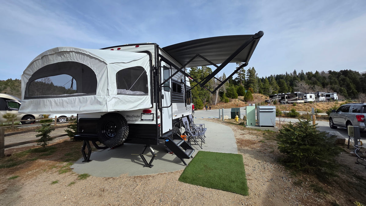 An RV rental, a travel trailer with a pop-out bed and awning, is set up at a scenic campsite among trees. — photo 13