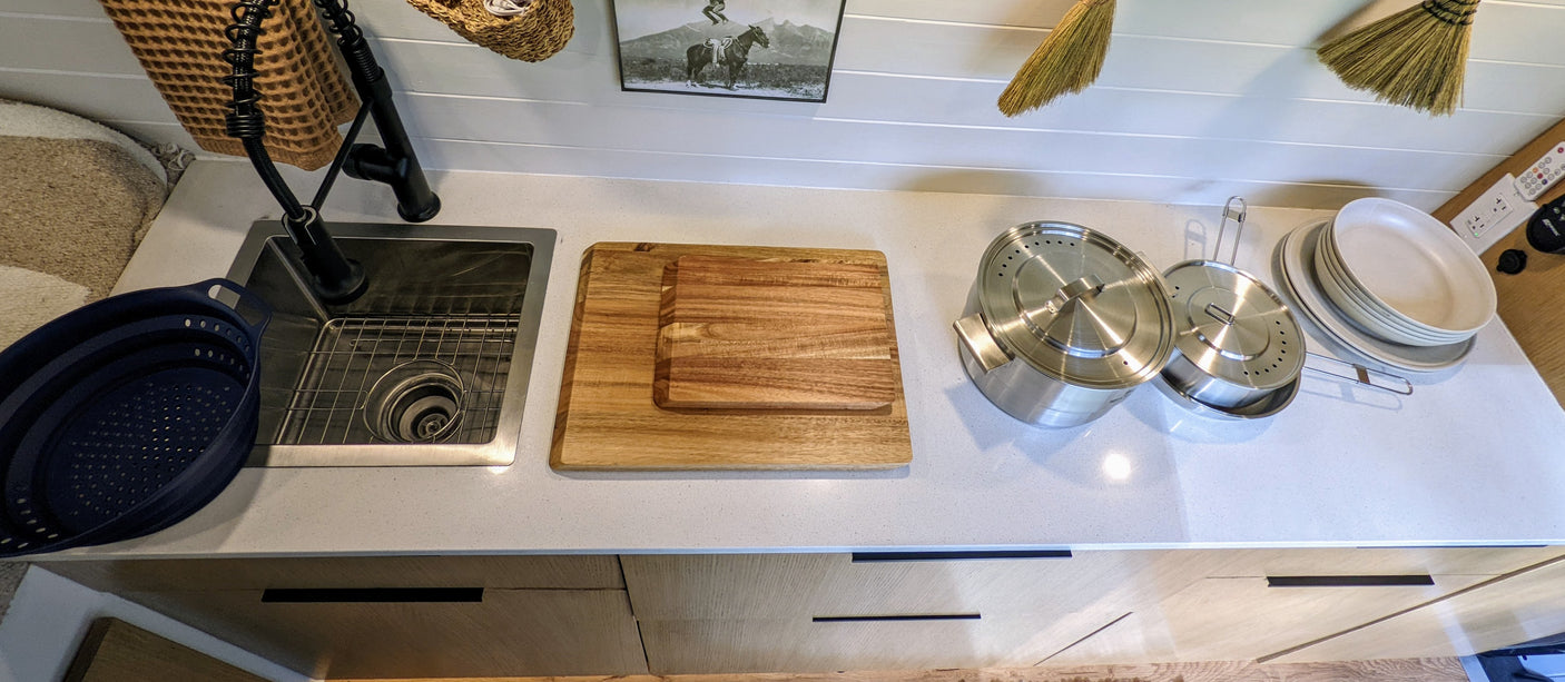 Organized RV rental kitchen counter: sink, faucet, collapsible colander, cutting boards, pots, and stacked dishes.