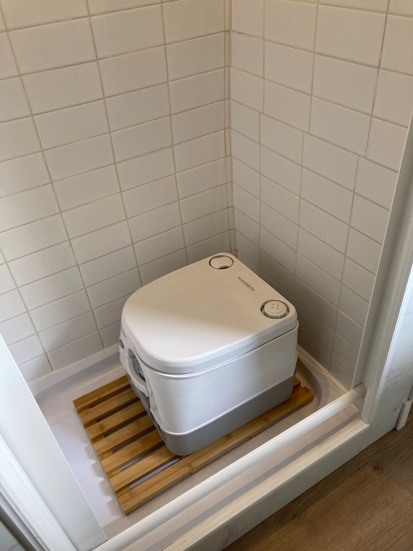 Compact Dometic portable toilet shown in a shower stall with white tiles and a wooden mat, suitable for an RV rental.
