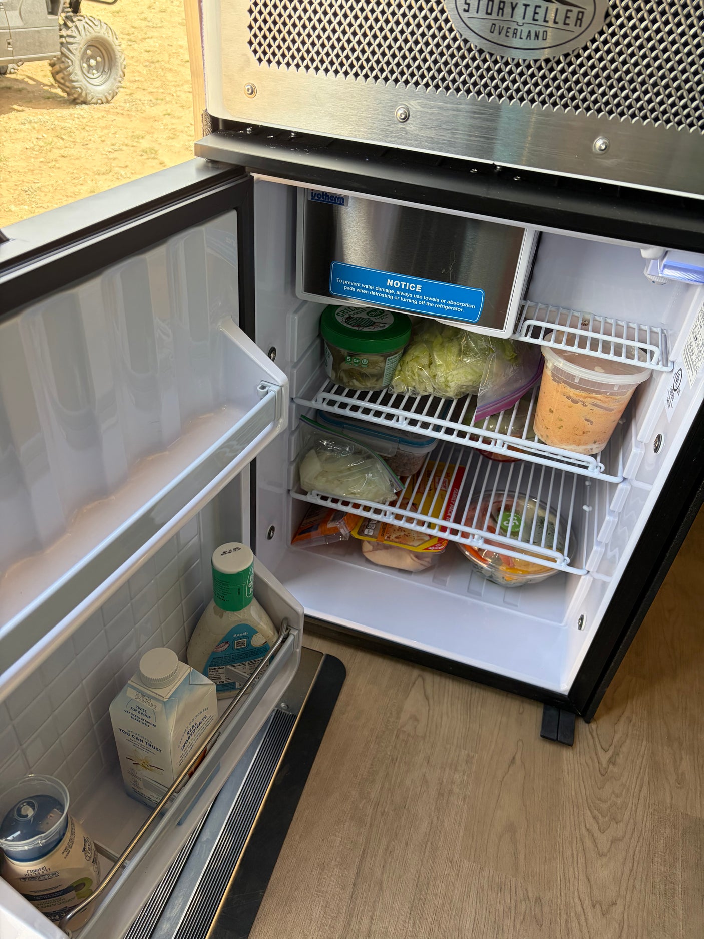 Open RV rental refrigerator full of food, drinks, and fresh produce, with a defrosting notice inside.