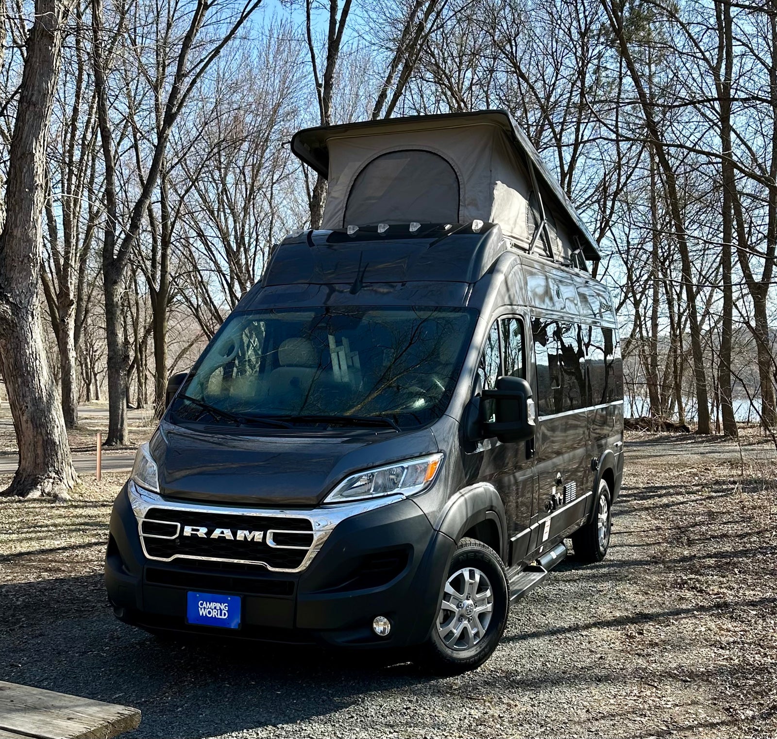 Sleek RV rental camper van with pop-top parked in a wooded campground near water. Ready for adventure!|A grey camper van (RV rental) with pop-top & bike rack is parked on gravel by a picnic table