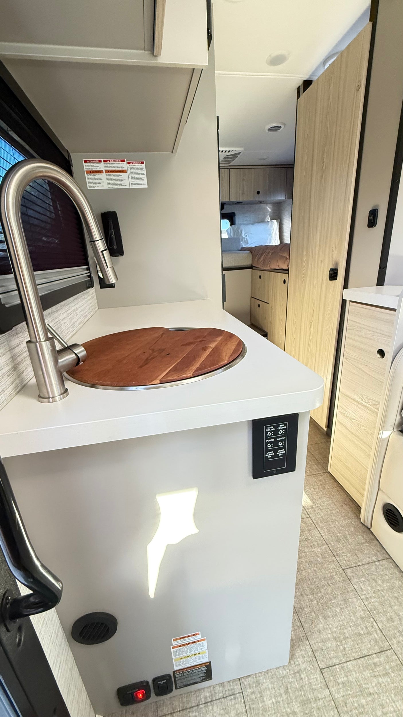 A bright, modern RV rental interior with a kitchen featuring a pull-down faucet, wooden sink cover, and controls. Beyond, a bed.