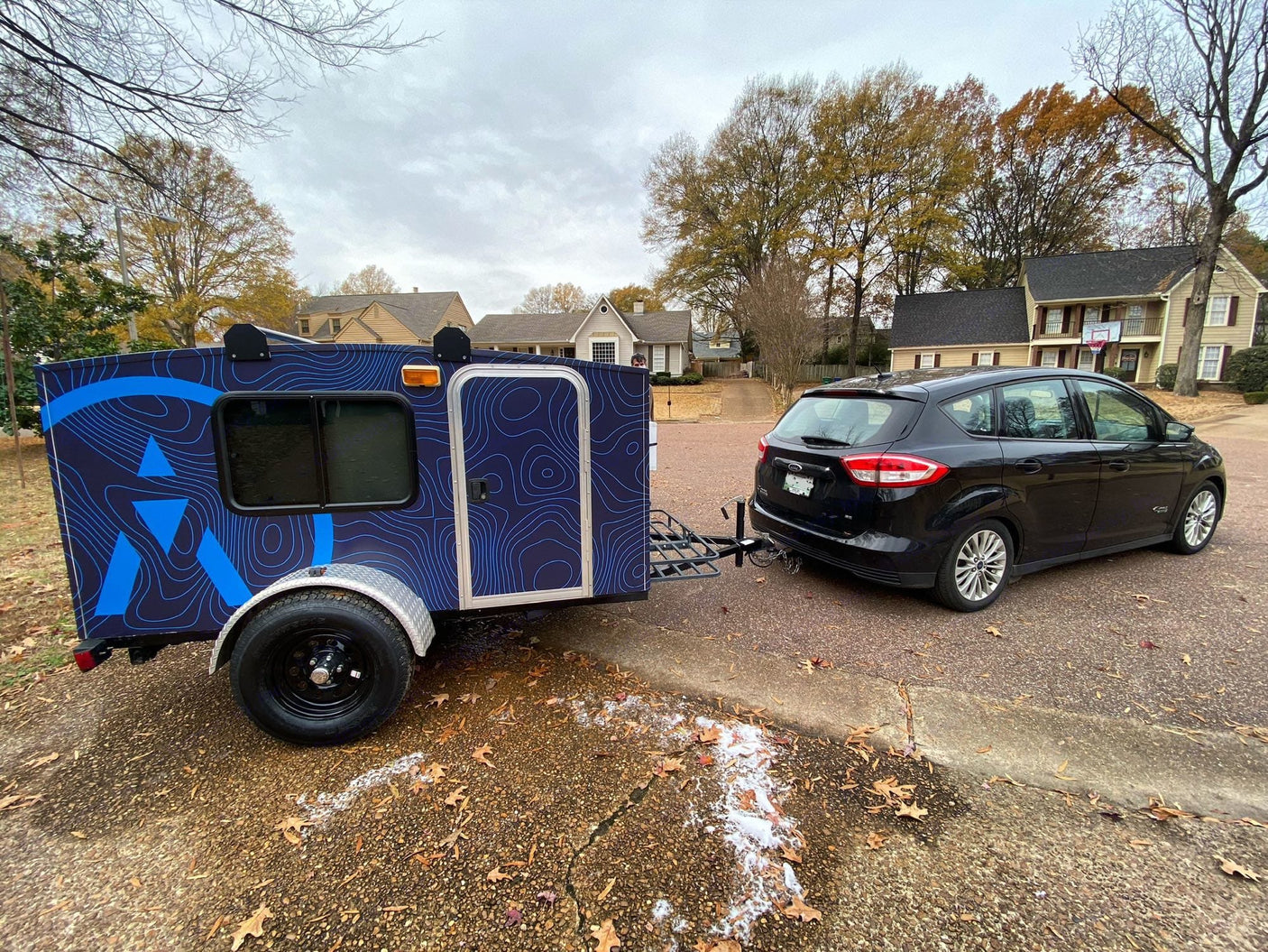 holiday-decorated RV rental camper sits on a lawn with festive lights. Nearby homes also sparkle brightly at night.|A unique blue teardrop RV rental is hitched to a black car
