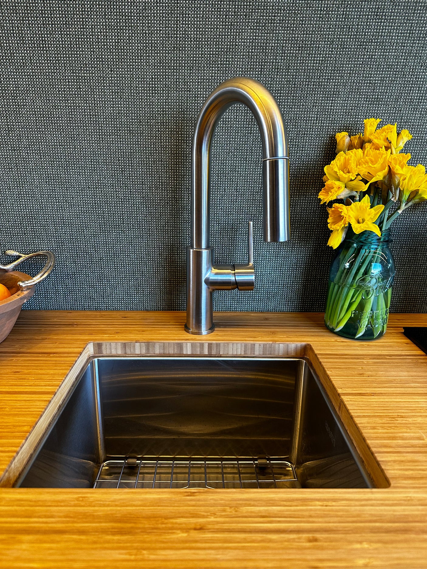 Sleek RV rental kitchen: bamboo counter, stainless sink with pull-down faucet, dark textured backsplash, and yellow flowers.