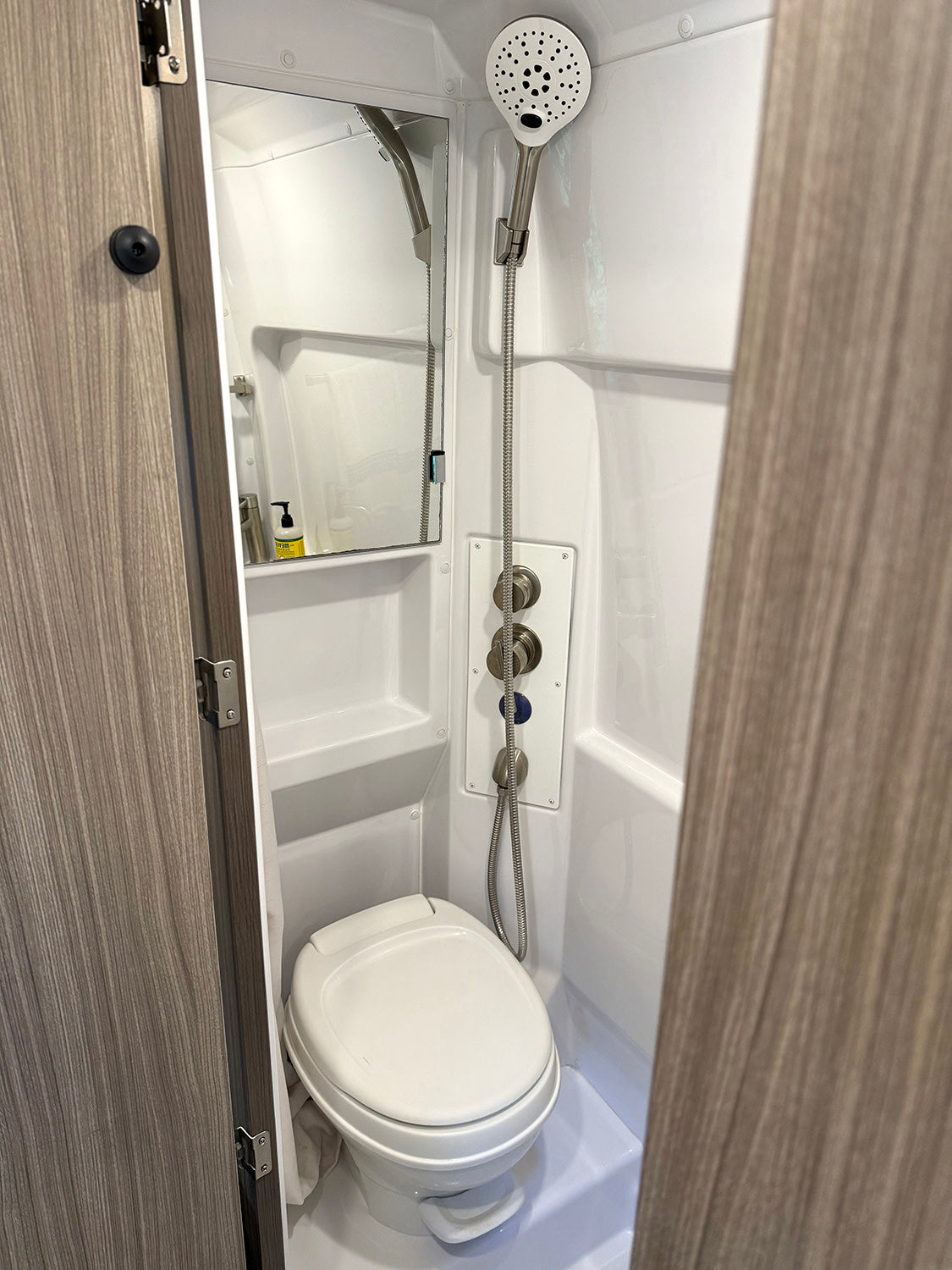 Experience the compact convenience of this RV rental's bathroom, featuring a white toilet, mirror, and handheld shower with controls.